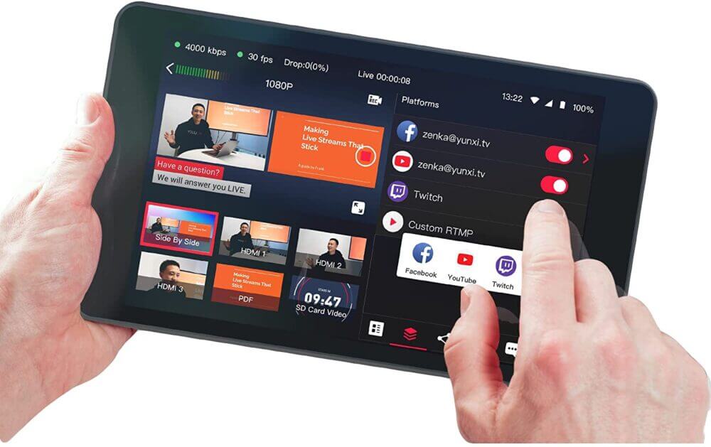  両手で保持されたタブレット型のライブ配信デバイス。画面左側に複数の入力ソースのプレビュー、右側にYouTubeやFacebook、Twitchなどの配信プラットフォームの選択メニューが表示される。
