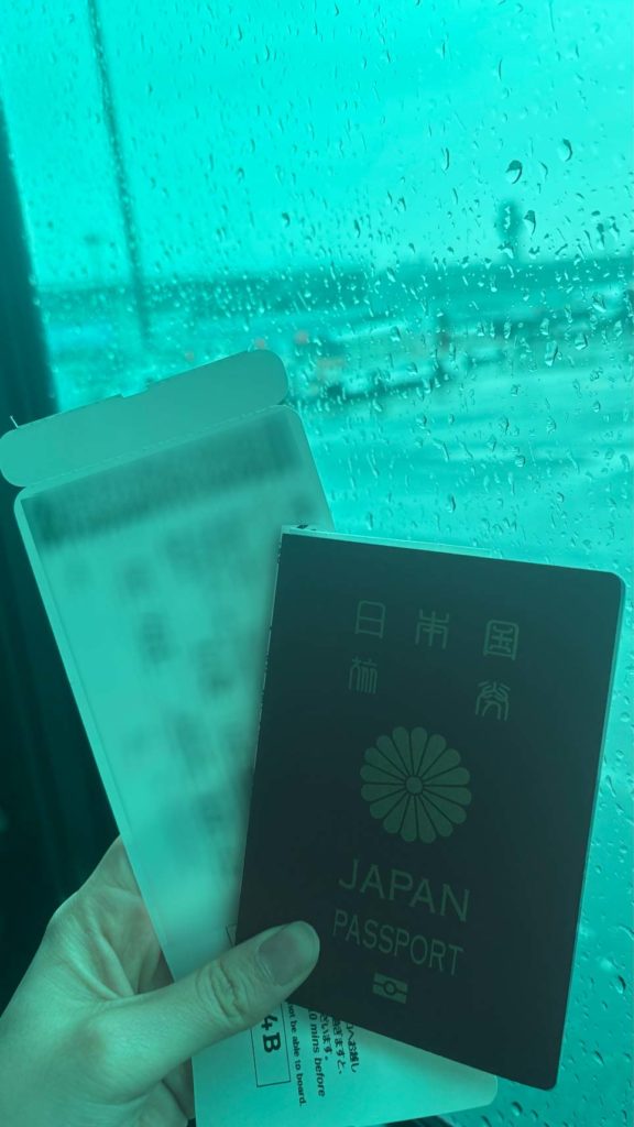 雨の日の空港の窓を背景に、手に持った日本のパスポートと深圳行きの航空券。