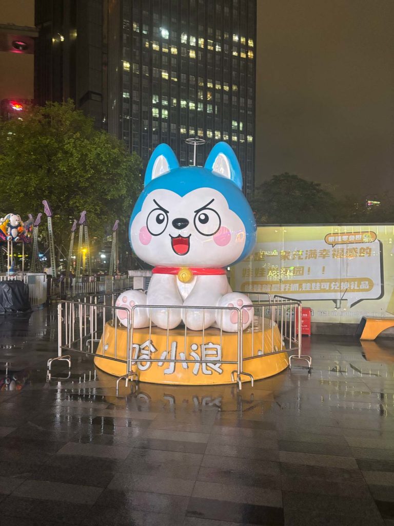 深圳の夜間の建物風景。複数の高層ビル群が照らされており、左側に赤い信号が見える。下部には青色のキャラクター（マスコット）の目がアップで映っている