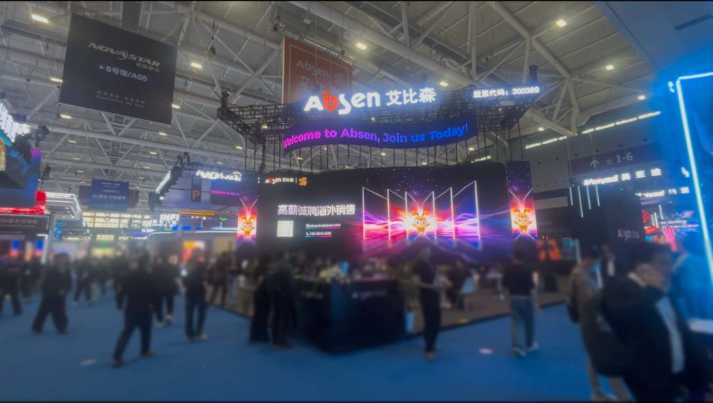 深圳のISLE会場。中央にAbsen(アビセン)社の巨大な黒い展示ブースがあり、鮮やかな高精細LEDディスプレイが光り輝いている。会場には青い絨毯が敷かれ、多くの来場者が行き交っている。
