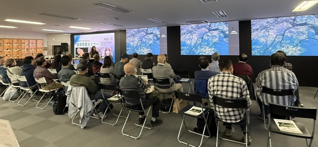 セミナー会場。モニターには桜の写真。講師が作品について話している。