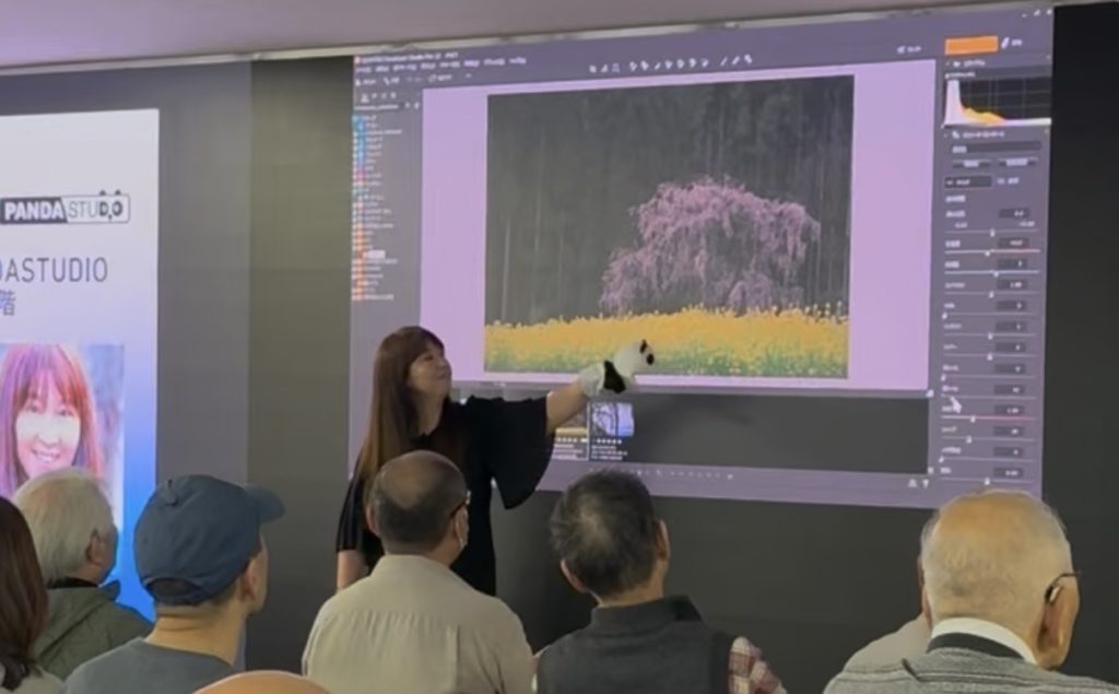 セミナーのモニターの前で説明する女性写真家。手にはパンダのぬいぐるみ。