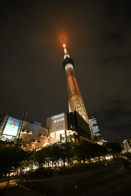 夜の東京スカイツリーを下から見上げた広角写真。タワーの鉄骨が白くライトアップされており、先端部分はオレンジ色に光って雲に少し隠れています。手前には「東京ソラマチ」や「すみだ水族館」のロゴが見える商業施設ビルがあり、ふもとには木々が植えられています